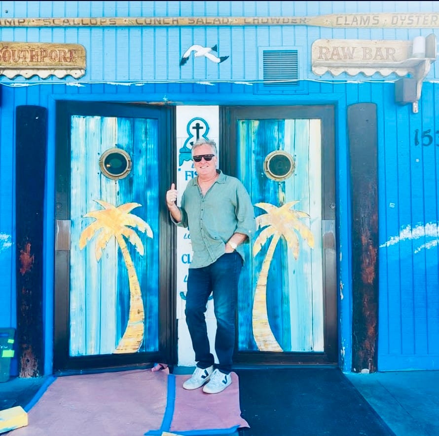 A man standing in the blue palm-tree doorway of Southport Raw Bar