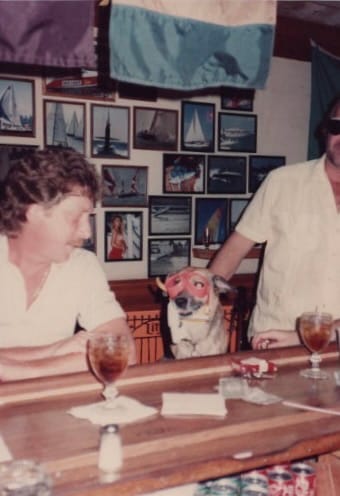 Two people and a dog at a table with drinks, in an old photo