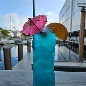 A tall blue cocktail with a pink paper umbrella and an orange slice on the dock rail, boats in the marina behind