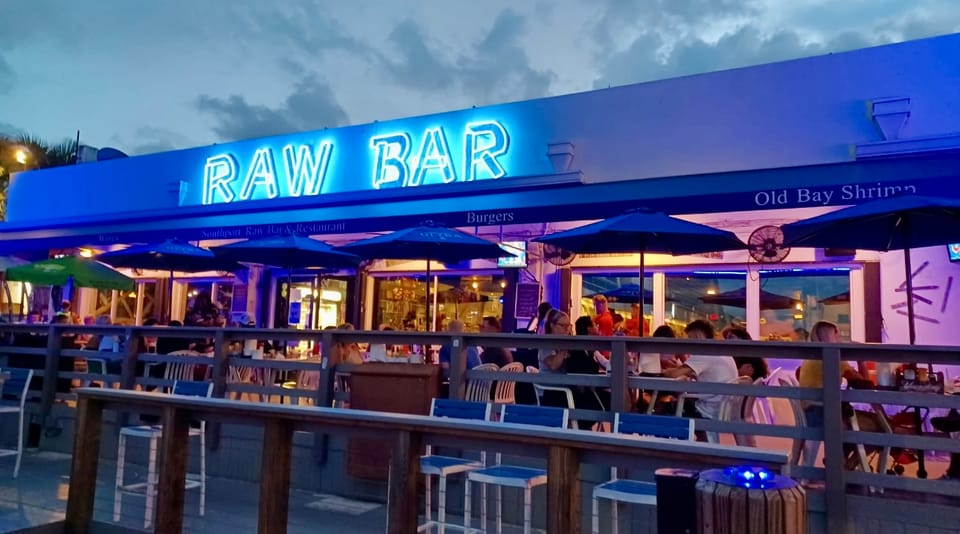 The RAW BAR sign in blue neon above the deck at dusk