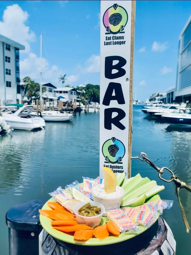 A platter of vegetables and dip on the dock rail beside the BAR sign, boats in the marina behind